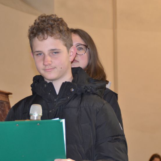 Ein junger Junge mit lockigem Haar hält ein Mikrofon und steht vor einer Frau mit Brille. Sie scheinen sich in einem Raum mit einer beigen Wand zu befinden.