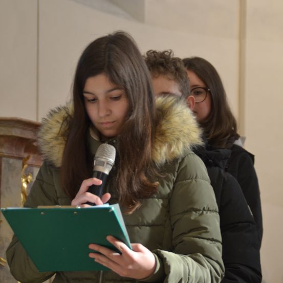 Ein Mädchen mit langen Haaren hält ein Mikrofon und schaut auf eine grüne Mappe. Sie trägt eine grüne Daunenjacke mit Pelzkragen. Hinter ihr stehen zwei Personen, eine mit Brille.