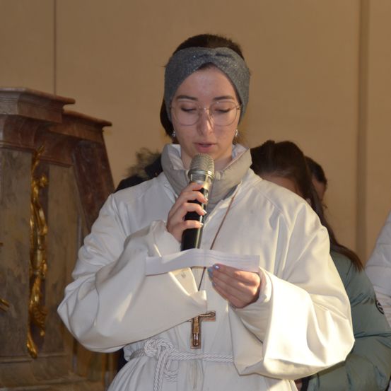 Eine Frau in einer weißen Robe hält ein Mikrofon und liest aus einem Papier in einer Kirche. Hinter ihr steht eine weitere Person. Der Hintergrund umfasst eine goldverzierte Holzstruktur.