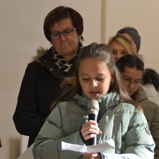 Ein junges Mädchen hält ein Mikrofon und liest von einem Stück Papier, mit Erwachsenen hinter ihr, einer mit Brille.