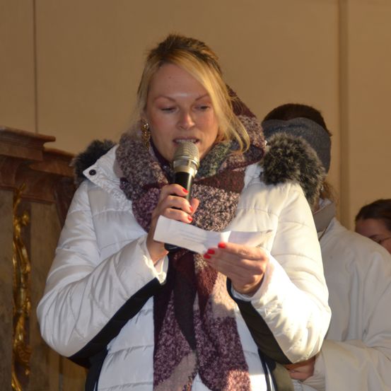 Eine Frau in einer weißen, gefütterten Jacke mit einem Pelzkragen liest von einem Zettel, während sie ein Mikrofon hält, und steht in einem Raum.