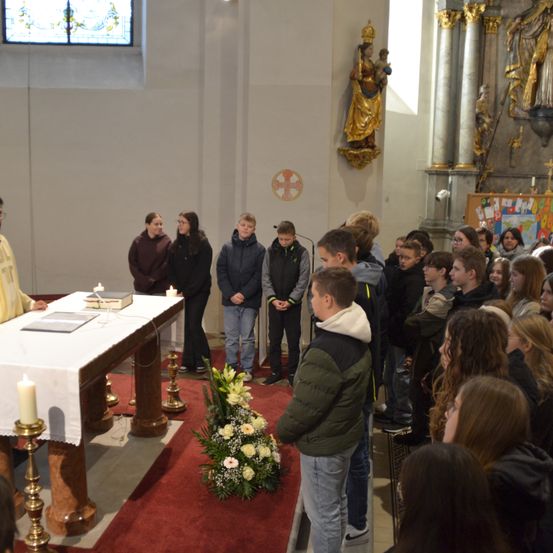 Eine Gruppe von Menschen steht in einer Kirche und sieht einem Altar mit Kerzen und Blumen zu. Sie scheinen einem sprechenden Priester zuzuhören.