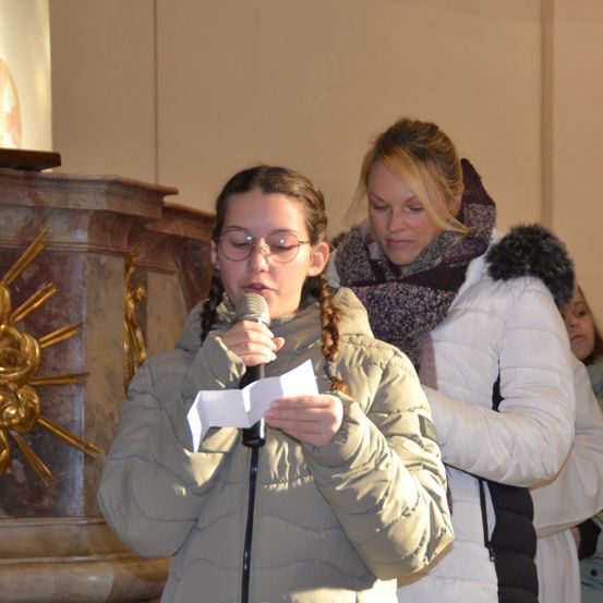 Ein Mädchen mit Brille und einer gefütterten Jacke liest aus einem Papier, während es ein Mikrofon in einer Kirche hält, mit einer Frau in einer mit Pelz gefütterten Jacke hinter ihr.