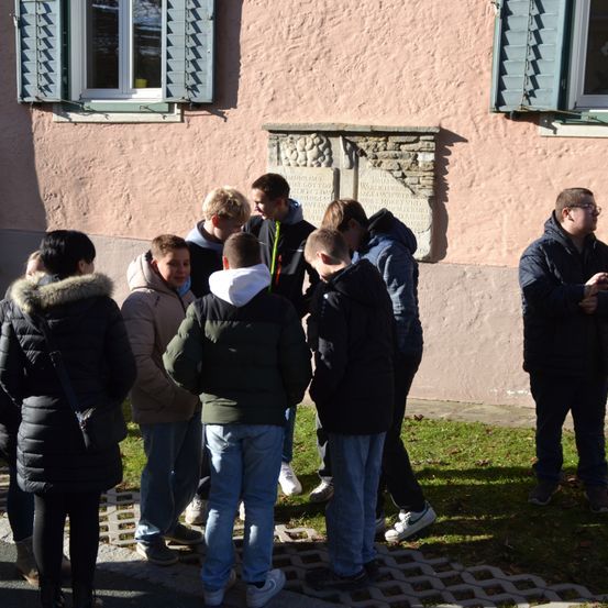 Eine Gruppe junger Männer und Frauen steht vor einem Gebäude mit einer Gedenktafel versammelt. Sie sind für kaltes Wetter gekleidet.