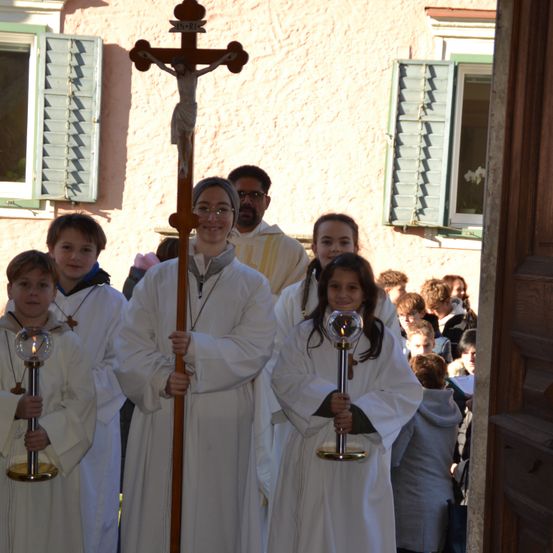 Eine Gruppe von Kindern in weißen Gewändern hält ein Kreuz und Kerzenleuchter in der Hand, während sie sich auf eine religiöse Zeremonie vor einem Gebäude mit grünen Fensterläden vorbereiten.