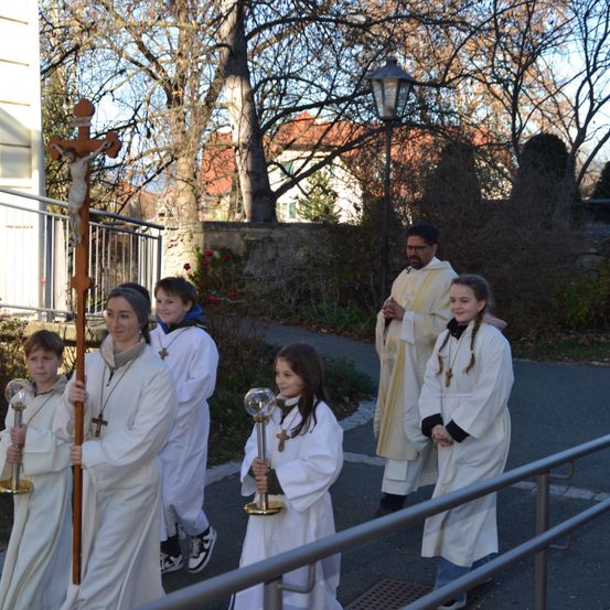 Eine Gruppe von Kindern und einem Priester gehen gemeinsam auf einem Pfad, halten ein Kreuz und eine goldene Kugel. Die Kinder tragen weiße Gewänder und Halsketten, und der Priester trägt ein gelbes Gewand. Sie sind von Bäumen und einer Straßenlaterne umgeben.