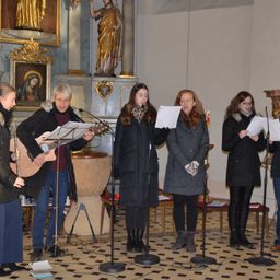 Eine Gruppe von Menschen singt in einer Kirche, eine Person spielt Gitarre und andere halten Papiere. Im Hintergrund steht eine Statue.