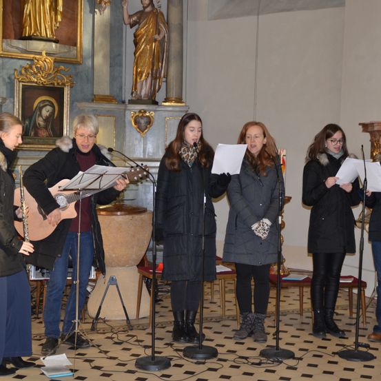 Eine Gruppe von Menschen singt in einer Kirche, eine Person spielt Gitarre und andere halten Papiere. Im Hintergrund steht eine Statue.