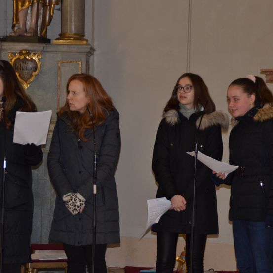 Vier Frauen stehen vor einem Mikrofon und halten Papiere in der Hand. Sie scheinen in einer Kirche zu singen. Hinter ihnen steht eine goldene Statue.