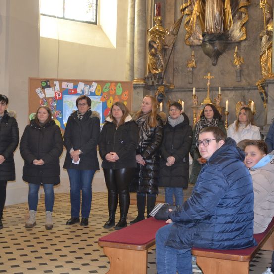 Eine Gruppe von Menschen, einige mit Brille, steht in einer Kirche, möglicherweise für eine Zeremonie. Sie sind in warme Kleidung gekleidet, und der Boden ist gefliest. Hinter ihnen befindet sich ein Bulletin Board.