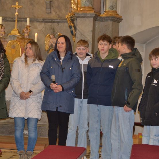 Eine Gruppe von Menschen steht in einer Kirche zusammen, eine Frau mit einem Mikrofon in der Hand und einem Kreuz im Hintergrund.