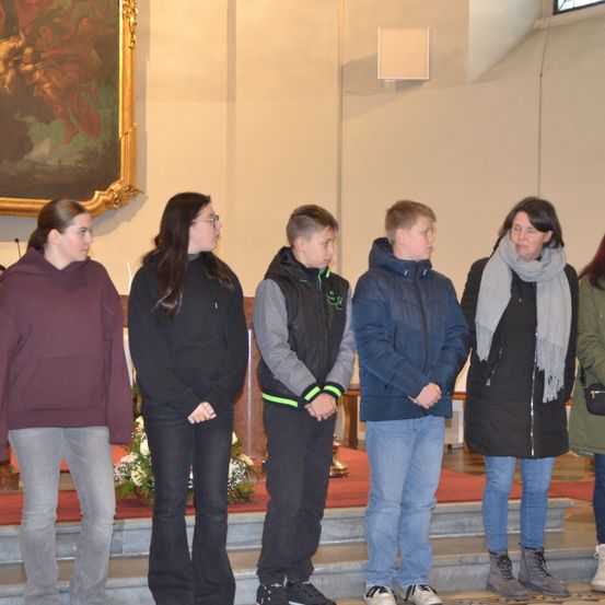 Sechs Personen stehen in einer Reihe in einer Kirche. Sie tragen alle Winterkleidung. Hinter ihnen befindet sich eine Blumendekoration auf dem Altar.