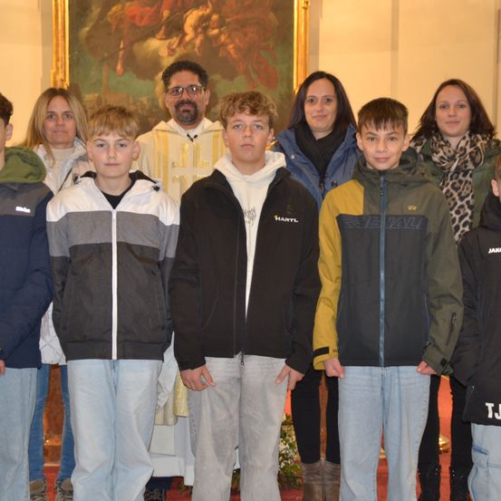Eine Gruppe von Jungen und Erwachsenen steht lächelnd in einer Kirche. Hinter ihnen steht ein Priester in einer weißen Robe. Die Wände sind mit einem großen Gemälde geschmückt.