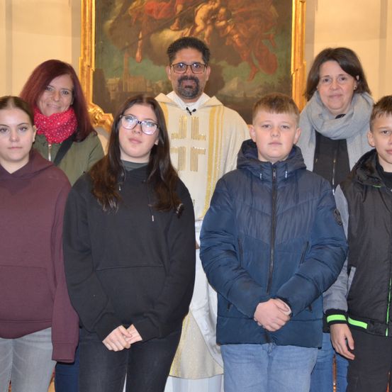 Ein Priester steht mit einer Gruppe junger Menschen und Erwachsener, lächelt vor einem religiösen Gemälde.