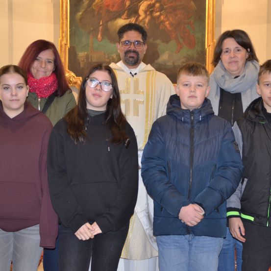 Ein Priester steht neben einer Familie vor einem Gemälde, alle warm gekleidet. Die Familie besteht aus einer Frau, einem Mädchen mit Brille und drei Jungen, von denen einer einen grünen Streifen an seiner Jacke hat.