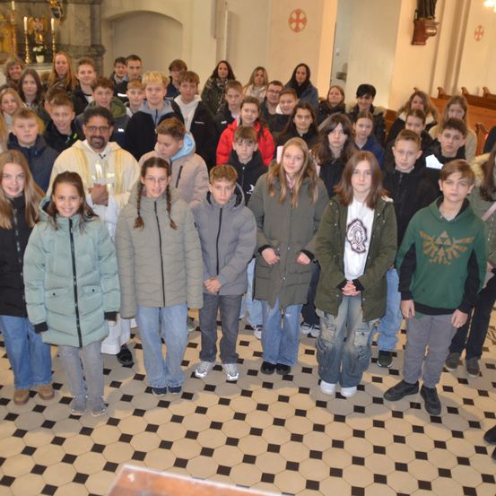 Eine große Gruppe von Menschen, darunter junge Männer und Frauen, steht in einer Kirche. Der Mann vorne trägt eine Brille und eine weiße Robe. Der Boden hat ein kariertes Muster.