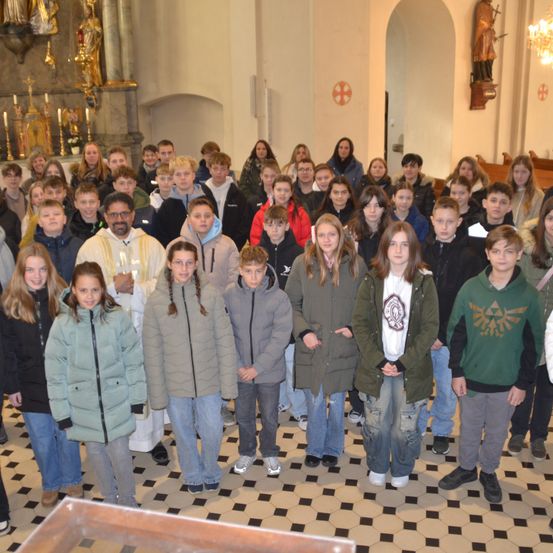 Eine Gruppe von Menschen ist in einer Kirche versammelt, mit einem Priester vorne. Einige tragen Jacken und Sneakers, während andere in Freizeitkleidung sind. Hinter ihnen ist der Altar mit Kerzen und einer Statue geschmückt.
