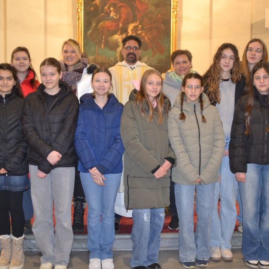 Eine Gruppe junger Menschen, wahrscheinlich Schüler, steht zusammen in einer Kirche. Ein Priester steht hinter ihnen. Die Kirche hat ein Gemälde an der Wand.
