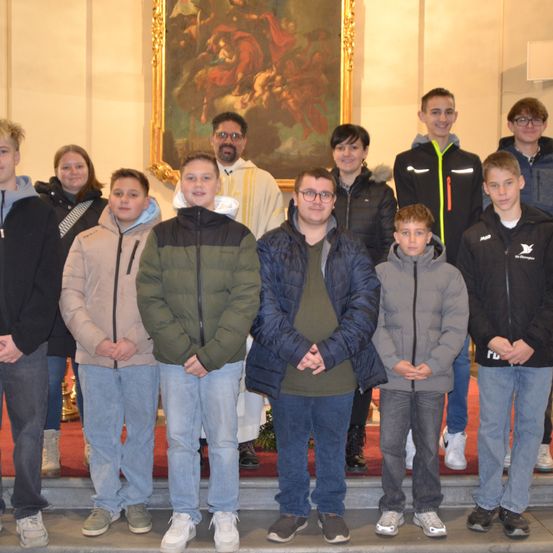 Eine Gruppe von Menschen posiert für ein Bild vor einem religiösen Gemälde. Es gibt zwei Erwachsene, einen Priester und mehrere Kinder, alle warm gekleidet in Jacken.