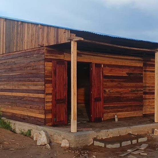 Ein Holzhaus mit einem grauen Metalldach und roten Türen steht auf einem Betonfundament. Es hat große Holzwände und eine offene Tür an einem bewölkten Tag.