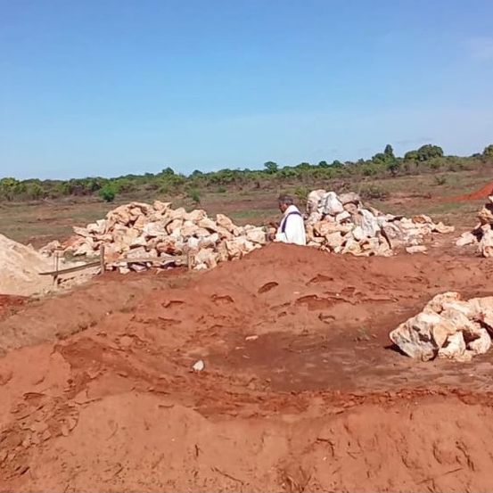 Ein Mann steht auf einem Feld mit Steinhaufen und Erde, unter einem klaren blauen Himmel. Bäume sind im Hintergrund zu sehen.