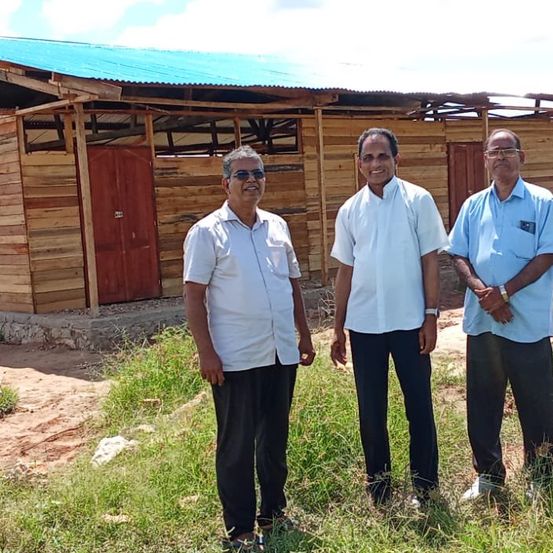 Drei Männer stehen vor einem Holzhaus mit blauem Dach. Sie sind in leichter Kleidung gekleidet, und die Umgebung ist im Freien mit Gras und klarem Himmel.