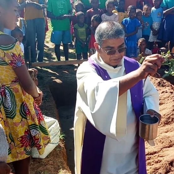 Ein Priester in einer weißen Robe schüttet Erde in ein Grab, während ein junges Mädchen in einem gelben Kleid in der Nähe steht. Eine Menschenmenge beobachtet.