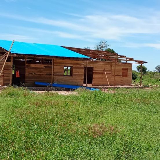 Ein Holzhaus im Bau mit einem blauen Dach steht mitten in einem Grasfeld. Eine Person steht in der Nähe des Eingangs.