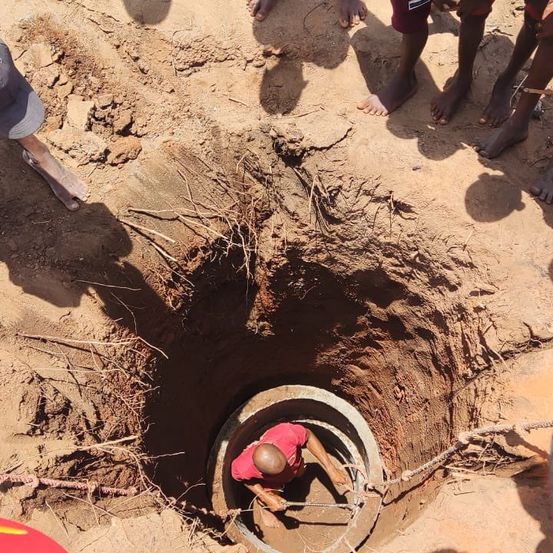Ein Mann in einem roten Shirt ist in einem Loch im Boden und arbeitet mit einer Kette. Mehrere Personen stehen umher, barfuß. Der Boden ist sandig und trocken.