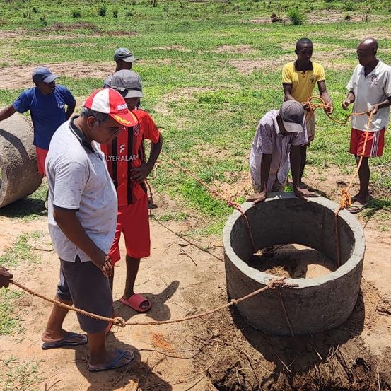 Eine Gruppe von Männern steht um einen großen Zementring auf einem Grasfeld. Einige tragen Hüte und Schuhe, andere sind barfuß. Sie benutzen Seile, um ein schweres Objekt zu ziehen.