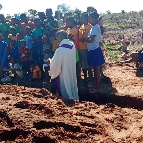 Ein Priester führt eine Beerdigungszeremonie in einem ländlichen Gebiet durch, umgeben von einer Gruppe von Menschen. Einige sitzen auf einer Holzbank, andere stehen, und ein Kind sitzt auf einem Baumstamm.