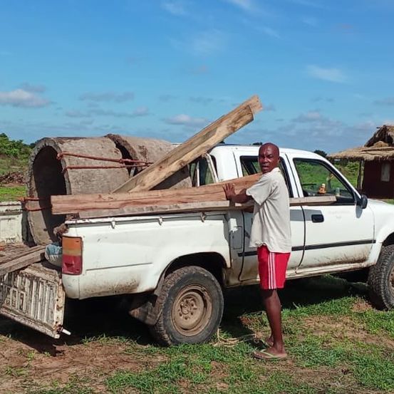 Ein Mann steht neben einem weißen Pickup-Truck, der mit Holzplanken beladen ist, in einem ländlichen Gebiet. Hinter ihm befindet sich eine kleine Hütte mit einem Strohdach.