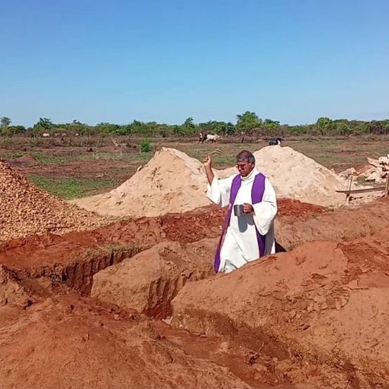 Ein Priester steht in einem Feld aus rotem Boden mit Steinhaufen und hält einen Becher in der Hand.