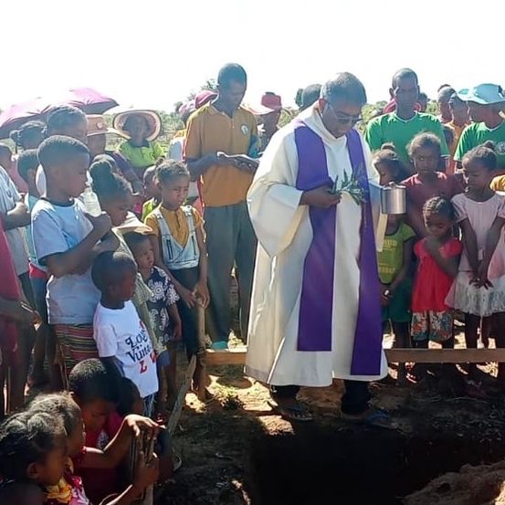 Ein Priester pflanzt einen Baum in ein flaches Loch, während eine Gruppe von Kindern und Erwachsenen zusieht. Der Priester trägt eine weiße und lila Robe und hält eine Pflanze.