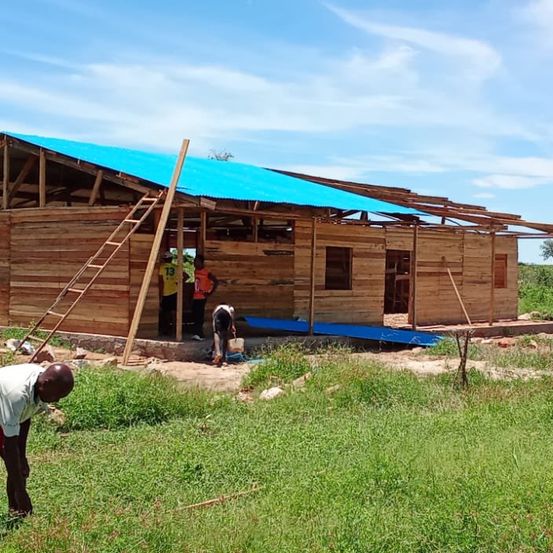 Unter einem blauen Tarp arbeiten mehrere Menschen an einem Holzhaus mit einem unfertigen Dach. Eine Leiter lehnt am Gebäude, und ein Mann beugt sich im Vordergrund.