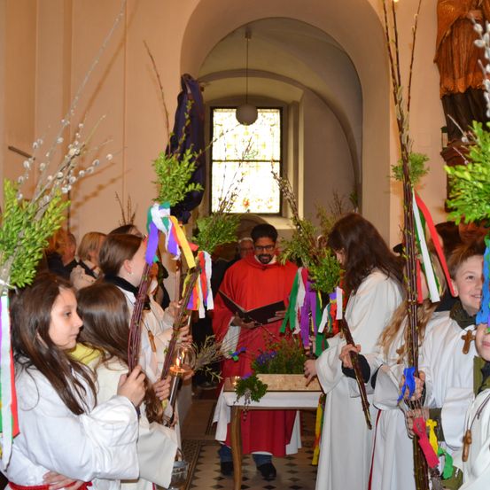 Eine Gruppe von Kindern mit Blumen und Kerzen nimmt an einem Gottesdienst teil. Ein Mann liest vor dem Altar aus einem Buch, umgeben von einem farbenfrohen Bogen.