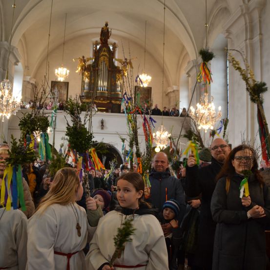 Menschen versammeln sich in einer Kirche, einige in weißen Gewändern, halten grüne Zweige. Ein Mann und eine Frau vorne tragen Brillen. Dahinter eine Menschenmenge mit bunten Bändern. Eine Orgel und Kronleuchter im Hintergrund.