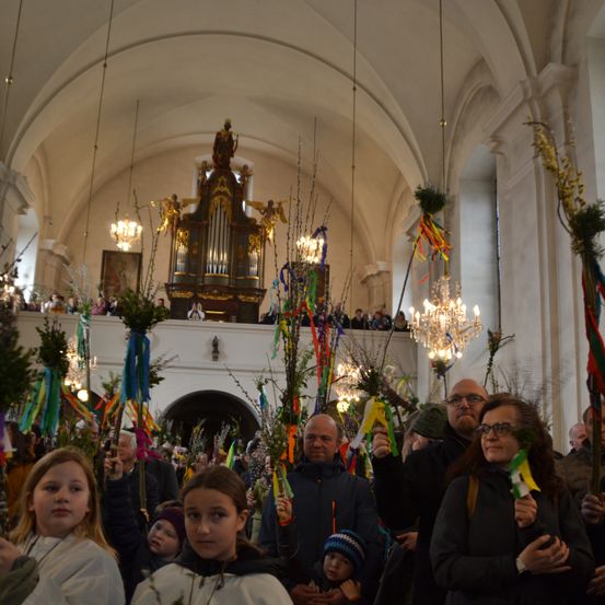 In einer Kirche versammeln sich Menschen mit geschmückten Zweigen in den Händen, während im Hintergrund Kronleuchter und eine Orgel stehen. Oben befindet sich eine Galerie mit Zuschauern.