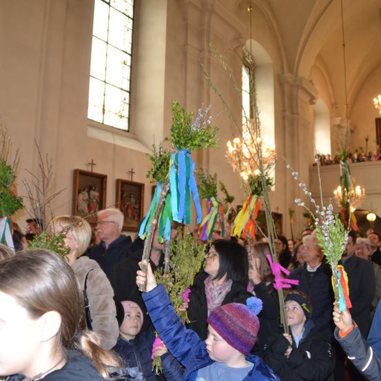 Eine Gruppe von Menschen, wahrscheinlich bei einem Kirchenereignis, hält dekorierte Zweige. Einige tragen Winterkleidung und Mützen, und der Raum ist gut beleuchtet.