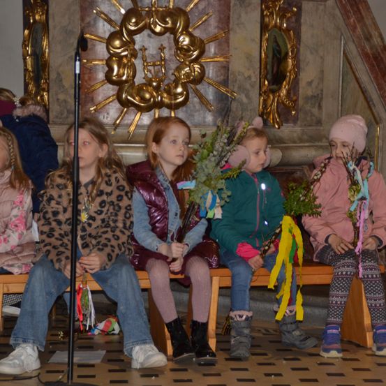 Fünf Mädchen sitzen auf einer Bank in einer Kirche, tragen Jacken und Stiefel. Sie halten Blumen und Bänder. Hinter ihnen ist eine goldene Sonne und ein Bilderrahmen an der Wand.