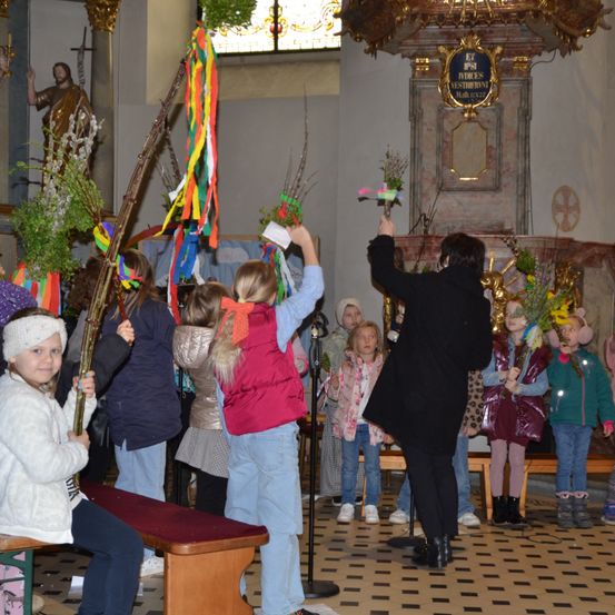 Kinder in Winterkleidung halten Palmzweige in der Kirche. Eine Frau mit Mikrofon steht vor ihnen. Eine Statue befindet sich links, eine Plakette rechts.