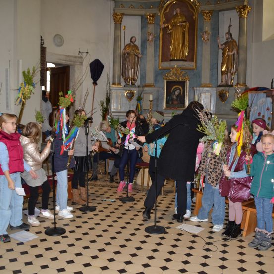 Kinder in einer Kirche, Blumen haltend und singend, mit einem Erwachsenen, der sie anführt. Eine Statue und ein Bildnis sind im Hintergrund.