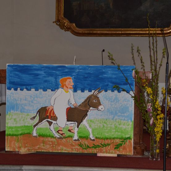 Ein Bild eines Mannes, der auf einem Esel auf einem grünen Feld reitet. Der Esel ist braun und weiß, und der Mann trägt eine weiße Robe und Sandalen.