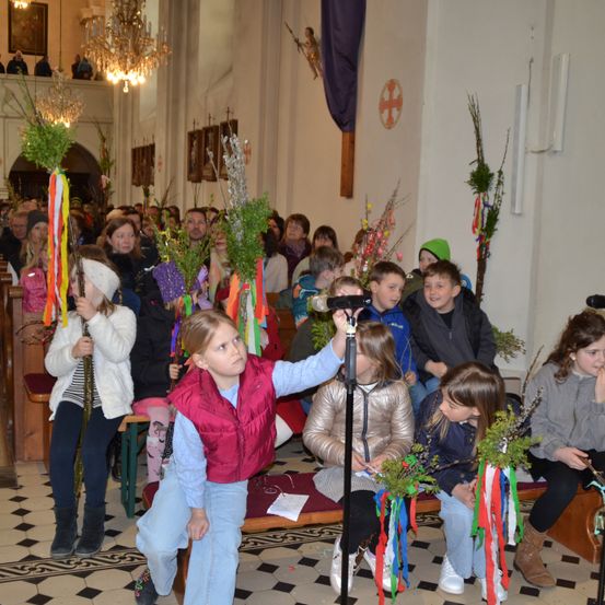 In einer Kirche halten Kinder bunte Bänder, während sie auf Bänken sitzen. Ein Mädchen mit einem Mikrofon in der Hand schaut nach oben. Hinter ihnen sitzen einige weitere Kinder, und einige stehen.