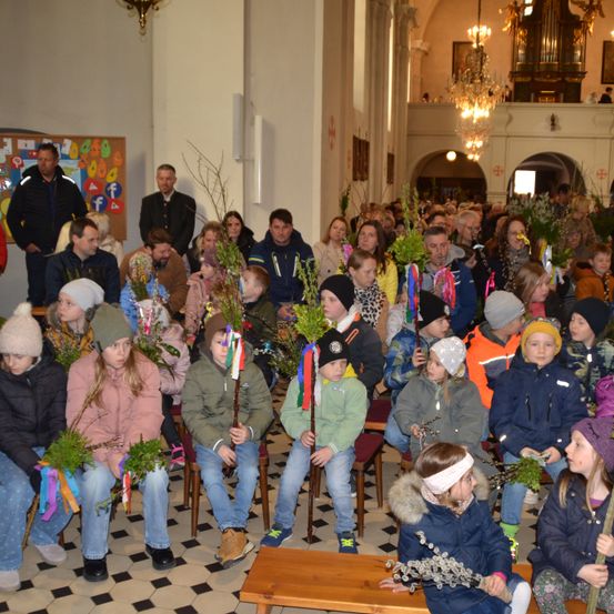 Eine Kirchenversammlung mit Teilnehmern, die Zweige und Stöcke halten. Die Menschen sitzen und stehen, und es gibt Kronleuchter und einen Aushang.