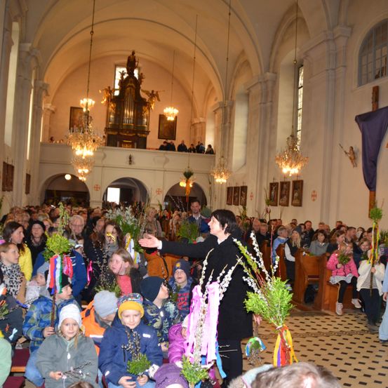 Eine Kirche voller Menschen, Kinder und Erwachsene, die Blumen halten und einem Mann in einem schwarzen Anzug zuhören. Der Raum hat Kronleuchter, eine große Orgel und Bänke.
