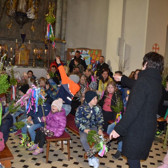 Eine Gruppe von Kindern und Erwachsenen sitzt auf Bänken in einer Kirche und hält dekorierte Pflanzen. Eine Person in einem schwarzen Mantel steht vor ihnen und leitet möglicherweise eine Zeremonie.
