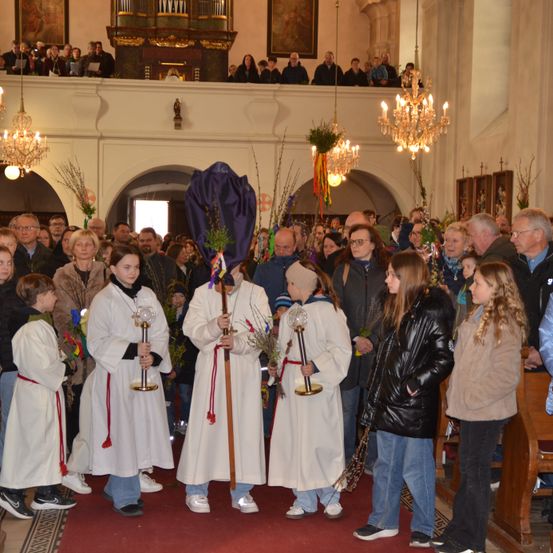 Mehrere Menschen stehen in einer Kirche und beobachten ein Ereignis. Drei Menschen in weißen Roben halten eine Kerze und einen Stab. Andere Menschen stehen an den Seiten und beobachten das Ereignis.