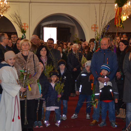 Eine Menschenmenge versammelt sich möglicherweise für eine Zeremonie in geschlossenen Räumen und hält Zweige. Ein Erwachsener in einer weißen Robe hält ein Kind mit Mütze. Einige tragen Winterkleidung.