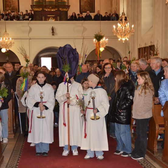 In einer Kirche laufen eine Gruppe von Menschen in weißen Gewändern und mit Stäben auf einem roten Teppich, umgeben von anderen Personen. Das Ambiente ist formal mit Kronleuchtern und Holzbänken.